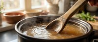 Wooden spoon stirring hot broth or soup in metal pot during cooking preparation in kitchen.