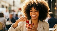 Young woman with curly afro hair and warm smile holding fresh raspberries outdoors in urban setting, enjoying healthy snack in city environment.