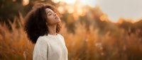 Young African American woman with curly hair enjoying peaceful moment in golden sunset light wearing cozy sweater in autumn field for wellness and mindfulness content.
