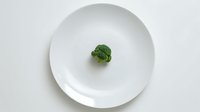 Fresh broccoli floret centered on round white plate against minimalist white background, highlighting healthy eating and portion control concept.