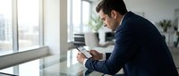 Young Caucasian businessman in navy suit using smartphone at glass desk in modern office, focused expression, natural window light.