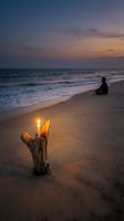 Peaceful beach sunset scene with person meditating by ocean waves and driftwood candle creating serene atmosphere for wellness and mindfulness concepts.