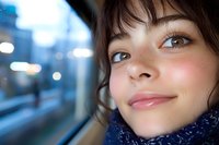 Young woman with gentle smile looking through window during evening commute, soft bokeh lights in background, wearing cozy blue scarf in winter.