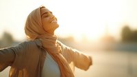 Young woman in hijab with arms outstretched enjoying sunset, expressing joy, freedom and spiritual connection in warm golden light.