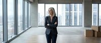 Middle-aged Caucasian female businesswoman in dark blazer standing in modern empty office space with floor-to-ceiling windows overlooking city skyline. Suitable for corporate, real estate, and business concepts.