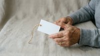 Senior man with weathered hands holding blank white gift tag with twine on neutral linen fabric background for holiday messaging and greeting card designs.
