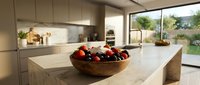 Wooden bowl filled with fresh red apples and dark grapes sits on light marble kitchen island in modern home with large windows overlooking garden landscape.