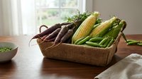 Woven basket filled with fresh organic vegetables including dark carrots, corn, and green beans on wooden table near window for cooking and meal preparation.