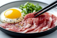 Fresh sliced raw pork belly with sunny side up egg and green onions on black plate, chopsticks picking up meat slice, close up view for Asian cuisine.