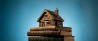 Miniature wooden house model sitting on stack of old books against blue background. Real estate education concept for mortgage learning and property investment.