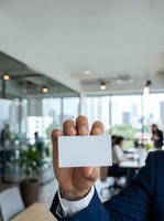 Male professional displaying blank white business card in modern office environment with colleagues working in background.