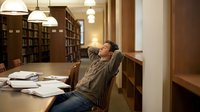 Young Asian male student stretching and relaxing at wooden desk in university library with books and warm lighting for study break concept.