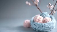 Soft pink yarn balls nestled in mint blue yarn nest against blurred background with delicate cherry blossom branch, creating gentle crafting composition.
