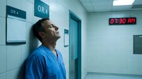 Exhausted male surgeon in blue scrubs leaning against hospital wall outside operating room after long night shift showing fatigue and stress.