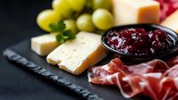 Appetizing charcuterie board with prosciutto, cheese selection, fresh grapes and cranberry sauce served on black slate plate. Gourmet food styling for restaurant menu.