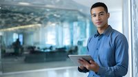 Young professional man in blue shirt holding digital tablet in modern office environment with blurred workspace background.
