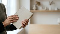 Person with medium skin tone holding open blank book indoors, wearing green cardigan. Use for education, reading, publishing, and learning concepts.