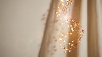 Warm glowing fairy lights on delicate wire branches against soft beige curtain, creating cozy bokeh ambiance for holiday and home decor.
