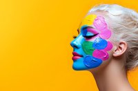 Side profile of blonde woman with creative multicolored face paint design featuring vibrant swirls in blue, pink, yellow and green against bright yellow background.