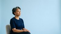 Mature woman with gray hair and dark skin wearing navy blue shirt sitting against light blue background, looking thoughtfully to side for portrait and corporate use.
