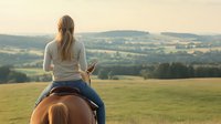 Young Caucasian woman with long blonde hair riding horseback on hilltop, viewing scenic rural landscape with rolling hills and meadows at sunset, back view.