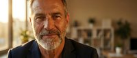 Mature man with gray beard and salt-and-pepper hair wearing dark blazer in modern office with warm natural light from window, suitable for corporate, business, and professional portraits.