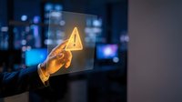 Businessman hand touching transparent screen with glowing yellow warning exclamation mark icon in modern office environment for cybersecurity alerts.
