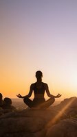 Woman practicing yoga meditation in lotus pose silhouetted against golden sunset sky on rocky cliff overlooking ocean for wellness and mindfulness.