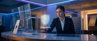 Young male professional in business attire working at modern desk with cybersecurity shield hologram and computer monitors in contemporary office environment for corporate technology concepts.