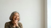 Senior woman with gray hair, wearing olive-toned sweater, smiling joyfully with hands clasped near face against white background. Suitable for healthcare, wellness, lifestyle and aging positively campaigns.