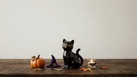 Black cat figurine with pumpkin, witch hat, and autumn leaves on wooden surface against neutral background for seasonal and holiday design projects.