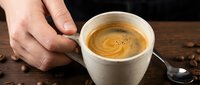 Caucasian male hand holding white ceramic cup of fresh espresso with crema on wooden surface with coffee beans and spoon.