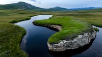 Meandering river curves through lush green grasslands with dramatic mountain backdrop, reflecting clouds in calm waters. Natural landscape shows erosion patterns.