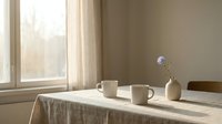 Minimalist dining table setup with two white ceramic mugs and small vase containing purple flower near bright window with natural sunlight streaming in.
