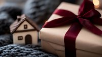 Elegant gift box with burgundy ribbon bow next to miniature wooden house on gray knitted texture, creating cozy holiday atmosphere with soft bokeh lights.