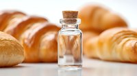 Small glass bottle with cork stopper against blurred background of fresh baked croissants and bread rolls, showcasing artisanal food ingredients.