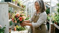 Mature woman with long gray hair and light skin tone, wearing a beige linen shirt and tan apron, arranging fresh coral and pink roses in a greenhouse filled with potted plants and wooden shelving.