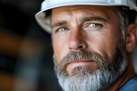 Mature construction worker with gray beard wearing white hard hat looking confidently at camera in industrial workplace setting for safety concepts.