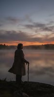 Silhouette of elderly man with walking stick standing on rocky shore at misty lake during golden hour sunrise reflecting on calm water surface.