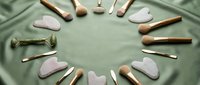 Rose quartz gua sha tools and facial massage accessories arranged in circle on sage green fabric background for skincare and wellness routine.