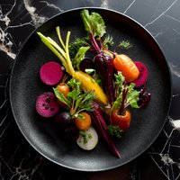 Fresh organic rainbow carrots and sliced beets arranged on black ceramic plate against dark marble background, garnished with herbs and greens.