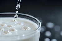 Fresh milk pouring into glass creating bubbles and drops on creamy white surface against dark background, macro shot with selective focus.