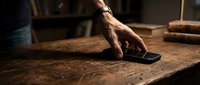 Man's hand reaching for smartphone on wooden desk in study room with books and warm lighting.