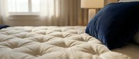 Comfortable bed with cream quilted mattress and navy blue pillow in bright bedroom with natural light from window and warm curtains.