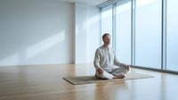 Middle-aged Caucasian man practicing meditation in lotus position on yoga mat in modern minimalist room with large windows and natural light.