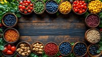Fresh berries, nuts, legumes and tomatoes in wooden bowls creating frame border on dark rustic background with green leaves and copy space for text.