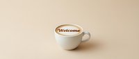 White ceramic cup of creamy latte with Welcome text on foam, placed on a warm beige background, evoking hospitality and comfort.