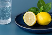 Fresh lemons on a dark blue ceramic plate with sparkling water glass and green leaves on a blue background.