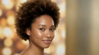 Young African American woman with natural curly afro hair smiling confidently with warm golden bokeh lights in background for beauty and lifestyle content.