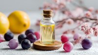 Glass bottle of essential oil on wooden coaster with fresh lemons and colorful grape berries against blurred cherry blossom background.